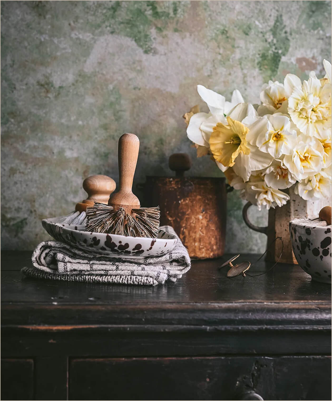 Wooden brushes in a speckled ceramic dish beside folded linens and a jug of daffodils
