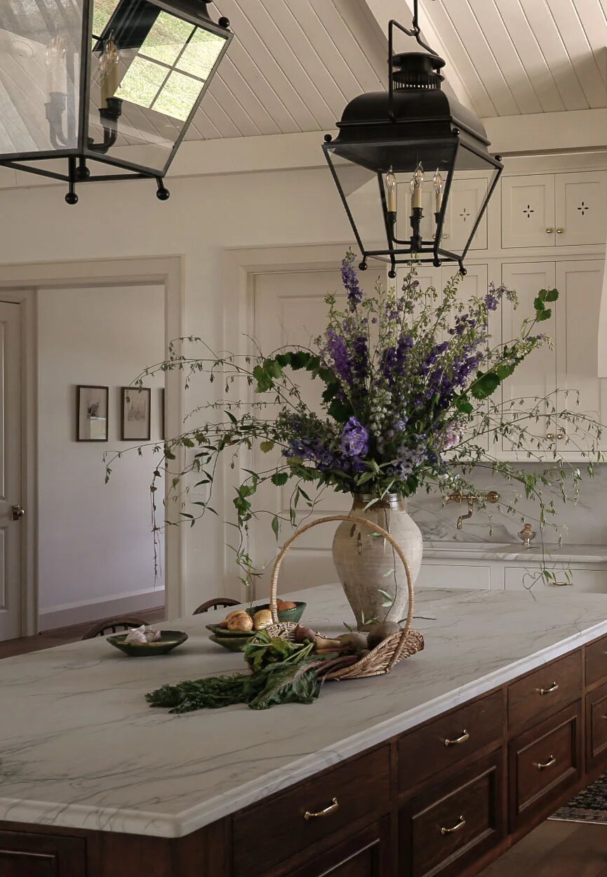 Ceramic vase of wild delphinium and greenery on a marble kitchen island under lanterns