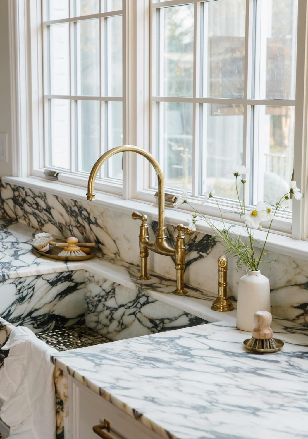 Marble kitchen sink with a brass bridge faucet beneath a sunny window